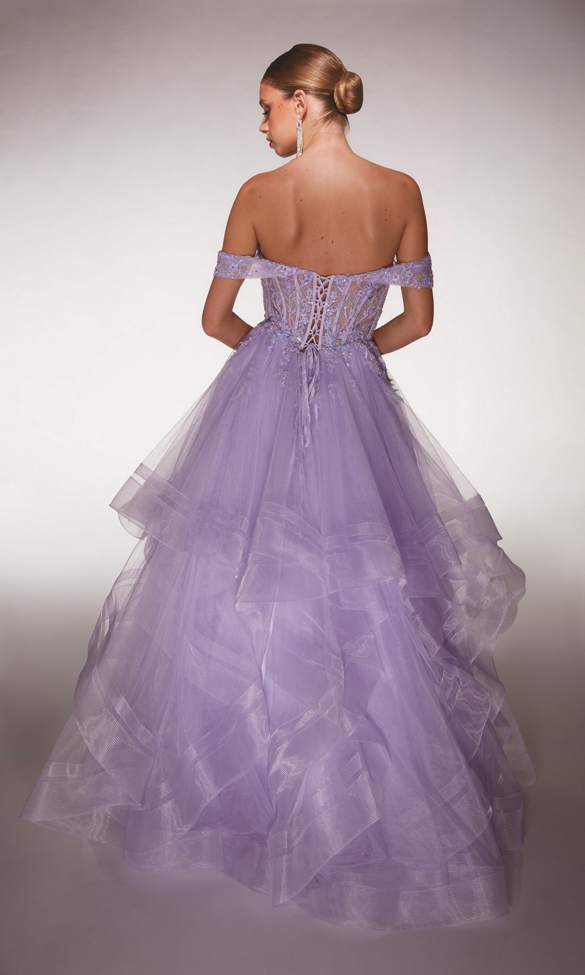 A woman with a low bun is wearing the Alyce 62207: a lavender off-the-shoulder tulle ball gown with lace-up back and floral lace appliqué, standing against a neutral background.